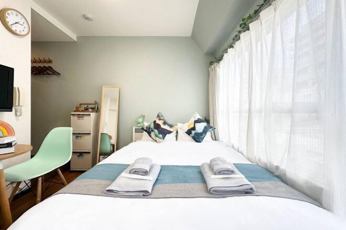 Habitación de hotel con cama, escritorio, silla, armario y ventana con cortinas translúcidas.