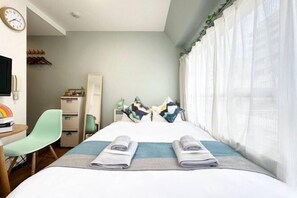 Habitación de hotel con cama, escritorio, silla, armario y ventana con cortinas translúcidas.