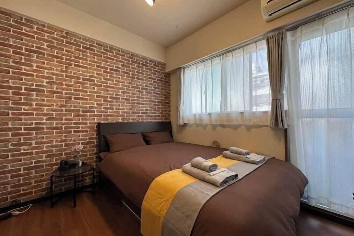 A hotel room with a brick wall, a large bed with brown bedding, a bedside table with a lamp, and a window with sheer curtains.