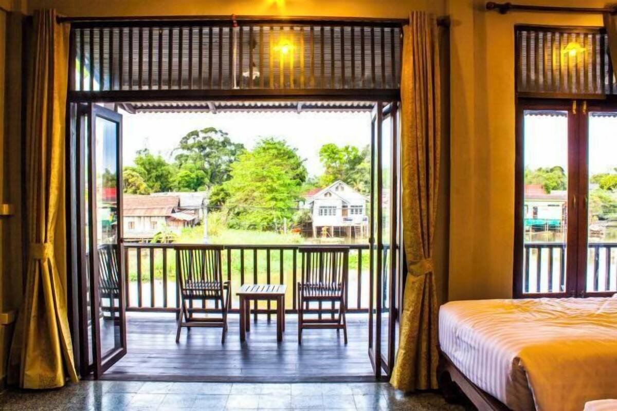 A room with a bed, a balcony with chairs and a table, and a view of greenery and buildings outside.