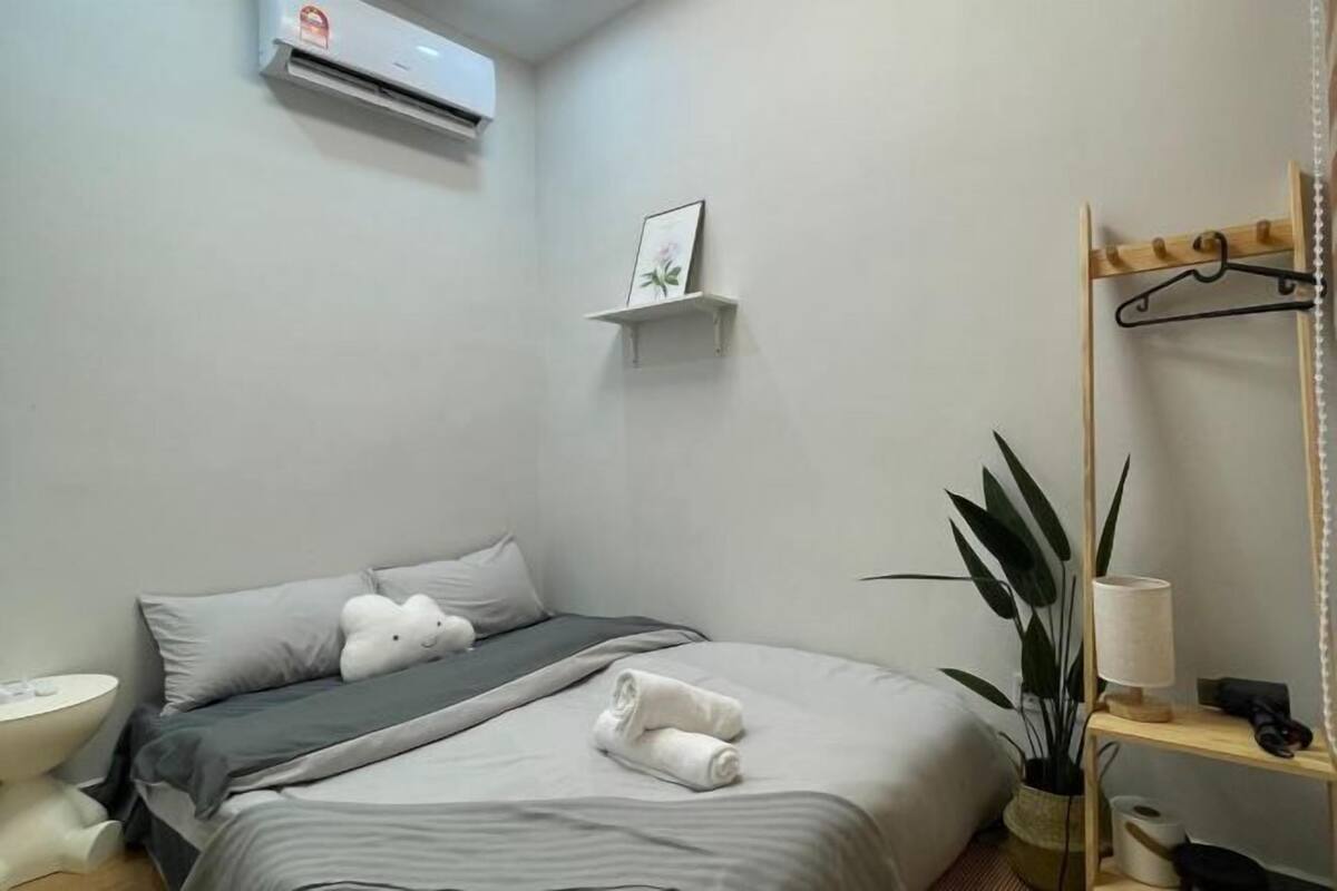 Une chambre d’hôtel bien rangée, avec un lit, une table de chevet, un climatiseur fixé au mur, une petite étagère avec un tableau, et un porte-manteau suspendu.
