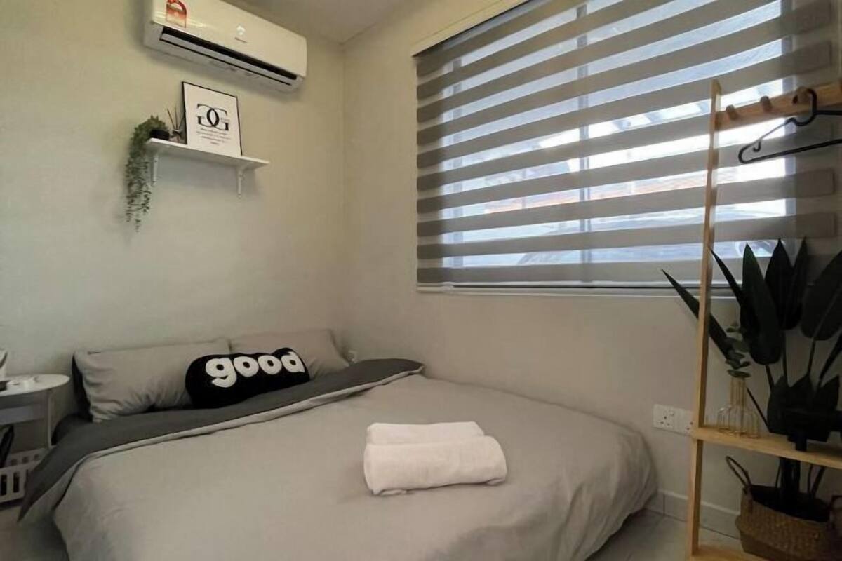 Une chambre bien rangée, avec un lit, une table de chevet, un climatiseur fixé au mur, une fenêtre avec des stores et une étagère ornée de plantes.