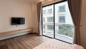 A hotel room with a bed, a TV mounted on the wall, a bench, a window with curtains, and a balcony with a railing. - Emiii House - Homestay Near The Wave Park Vinhomes Ocean Park 2 (Nghia Tru)