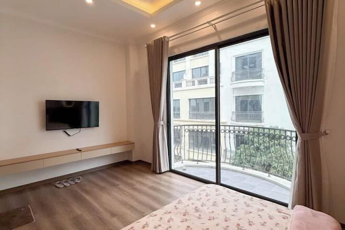 Une chambre d’hôtel avec un lit, un téléviseur fixé au mur, un banc, une fenêtre avec des rideaux et un balcon avec une balustrade.