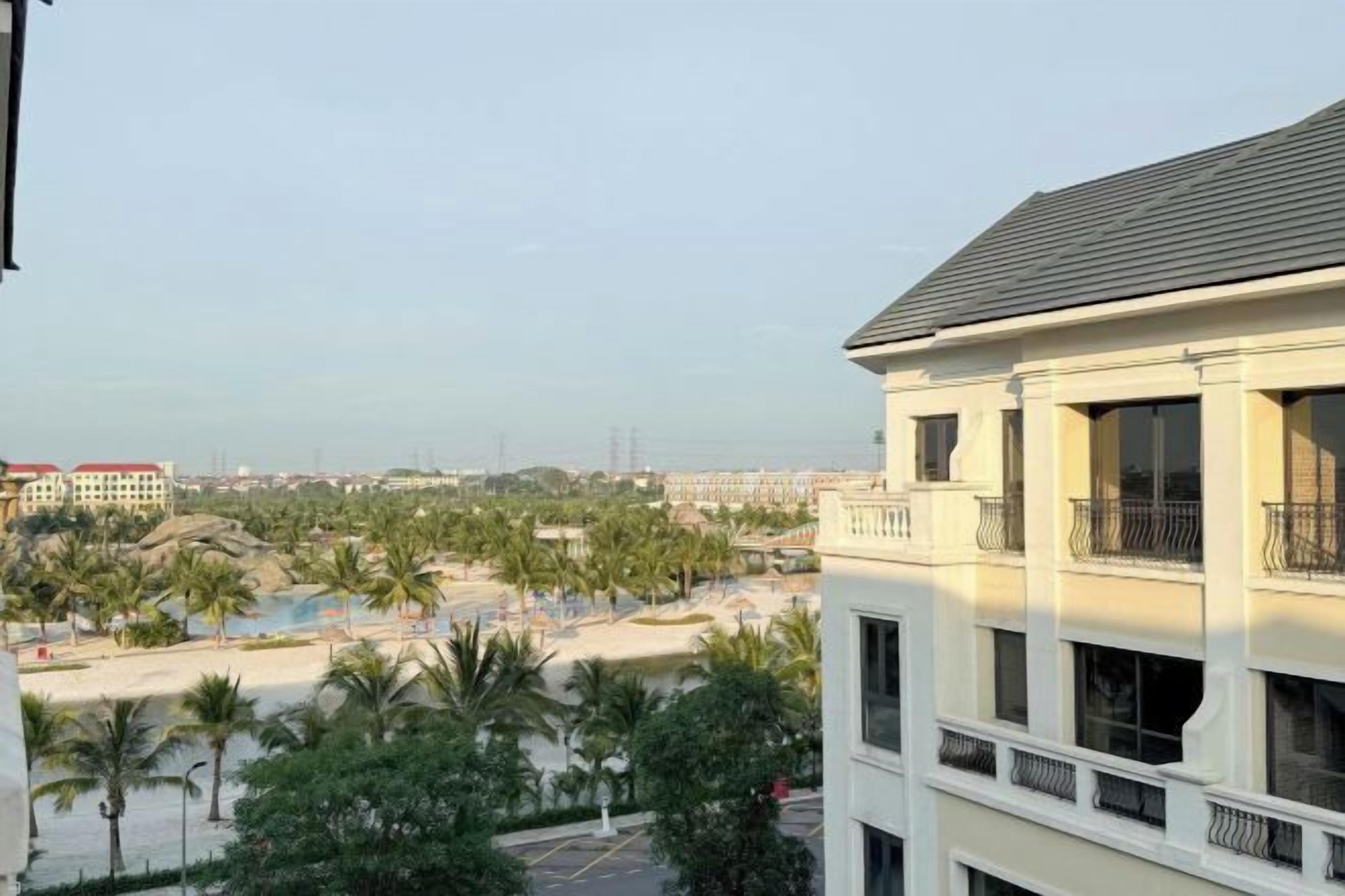 A multi-story building with balconies, a pool area with palm trees, and a view of a beach and distant buildings.