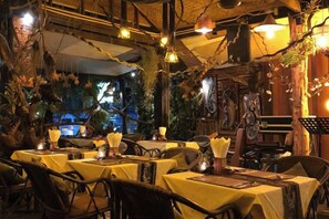 Área de refeições interna com mesas preparadas para hóspedes, com decoração rústica, luminárias pendentes e plantas.