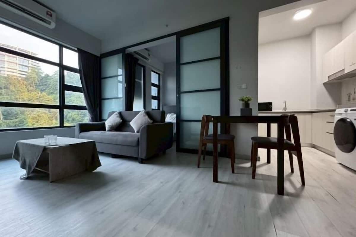 A modern living room with a grey sofa, a wooden coffee table, and a dining area with a wooden chair. There is a kitchen with a washing machine and a window with curtains.