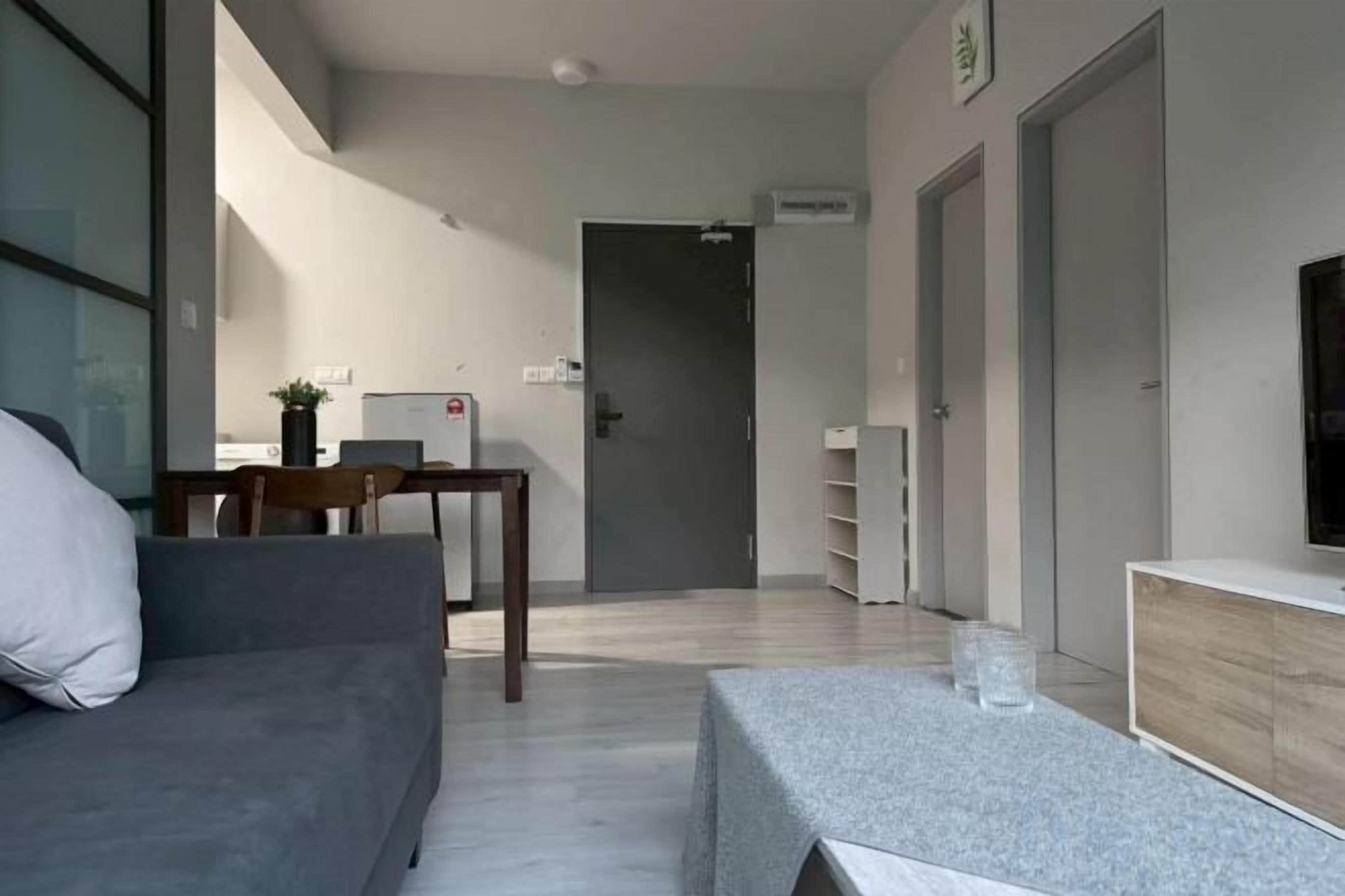 Un salon moderne avec un canapé, une table basse et un téléviseur. On aperçoit à l’arrière-plan un coin cuisine avec un réfrigérateur et une table à manger.