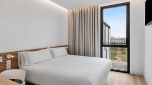 A modern hotel room with a large bed, a desk, and a view of the city through a large window.