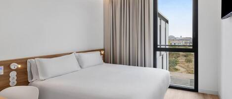 A modern hotel room with a large bed, a desk, and a view of the city through a large window.