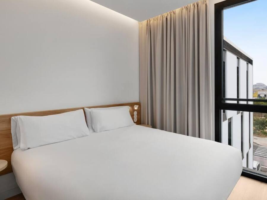 Ein Hotelzimmer mit einem großen Bett, einem Nachttisch, einem Fenster mit Vorhängen und Blick ins Freie.