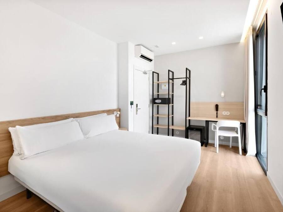 A modern hotel room with a large bed, a desk, a chair, and a wardrobe.