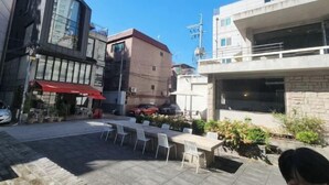 Un área de estar al aire libre con mesas y sillas, un toldo rojo y un edificio de grandes ventanales y fachada de ladrillo.