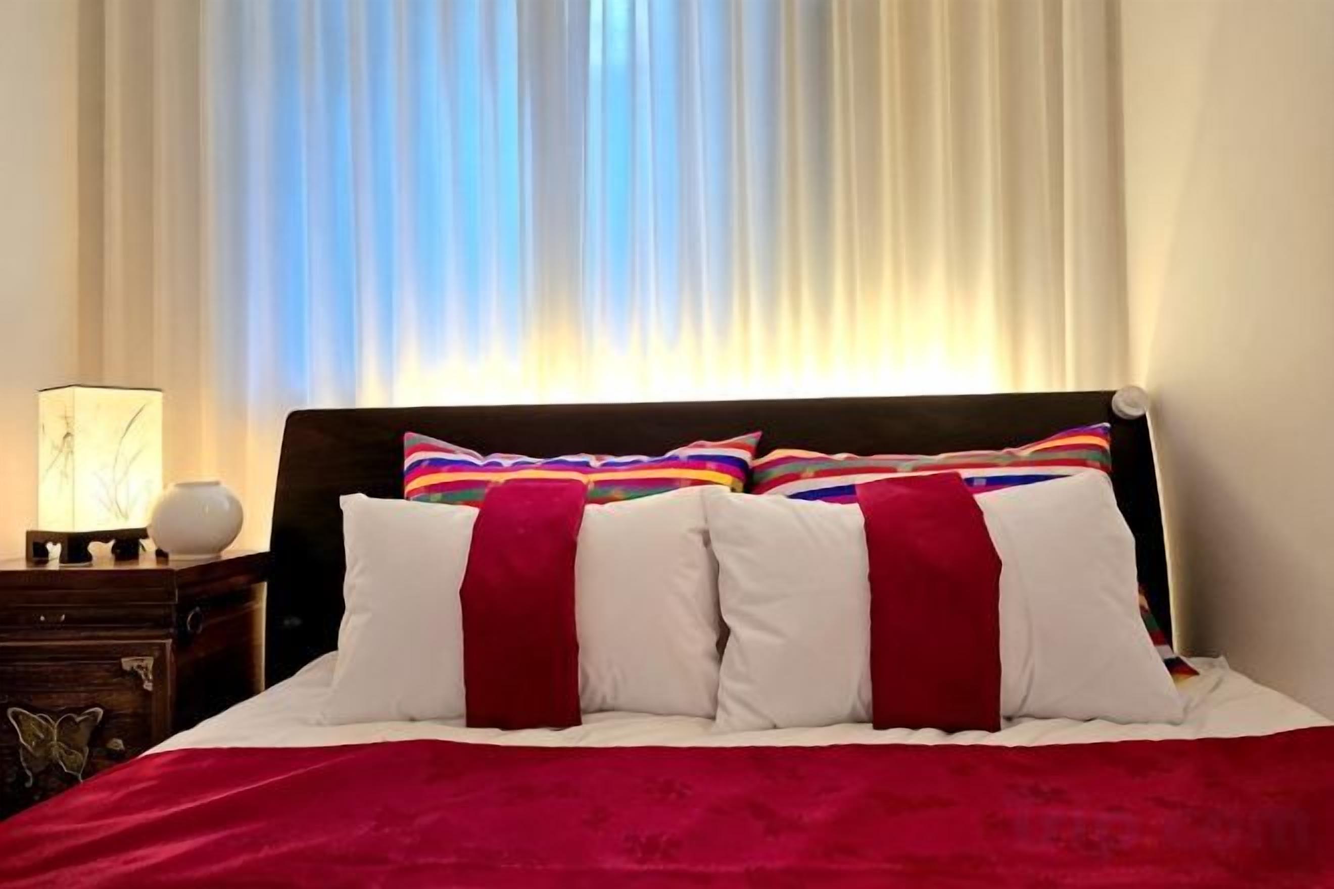 Una cama con ropa de cama blanca y roja, un cabecero de madera y una mesita de noche con una lámpara y un florero.