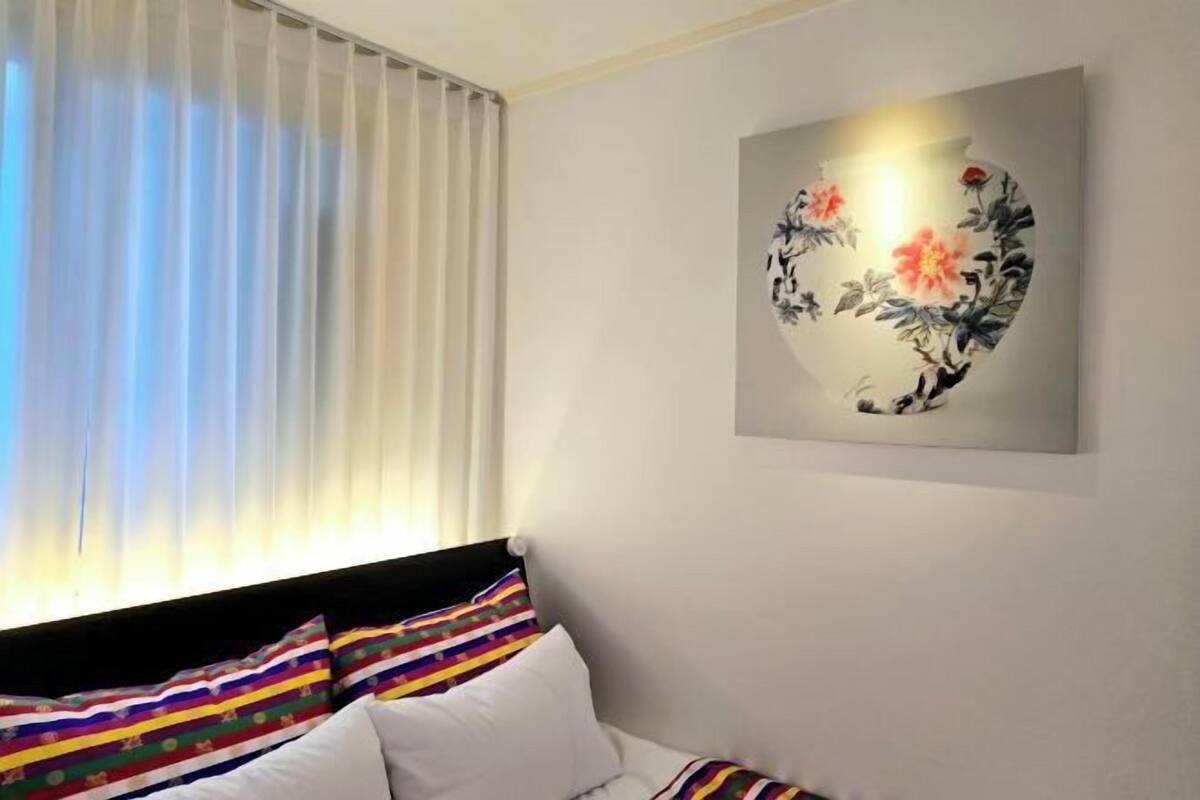 Ein Hotelzimmer mit einem Bett, farbenfrohen Kissenstreifen, einem Fenster mit transparenten Vorhängen und einem an der Wand hängenden Blumenbild.