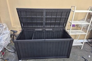Un meuble de rangement noir avec un couvercle à charnières, un vélo et une étagère en métal blanche, le tout adossé à un mur beige.