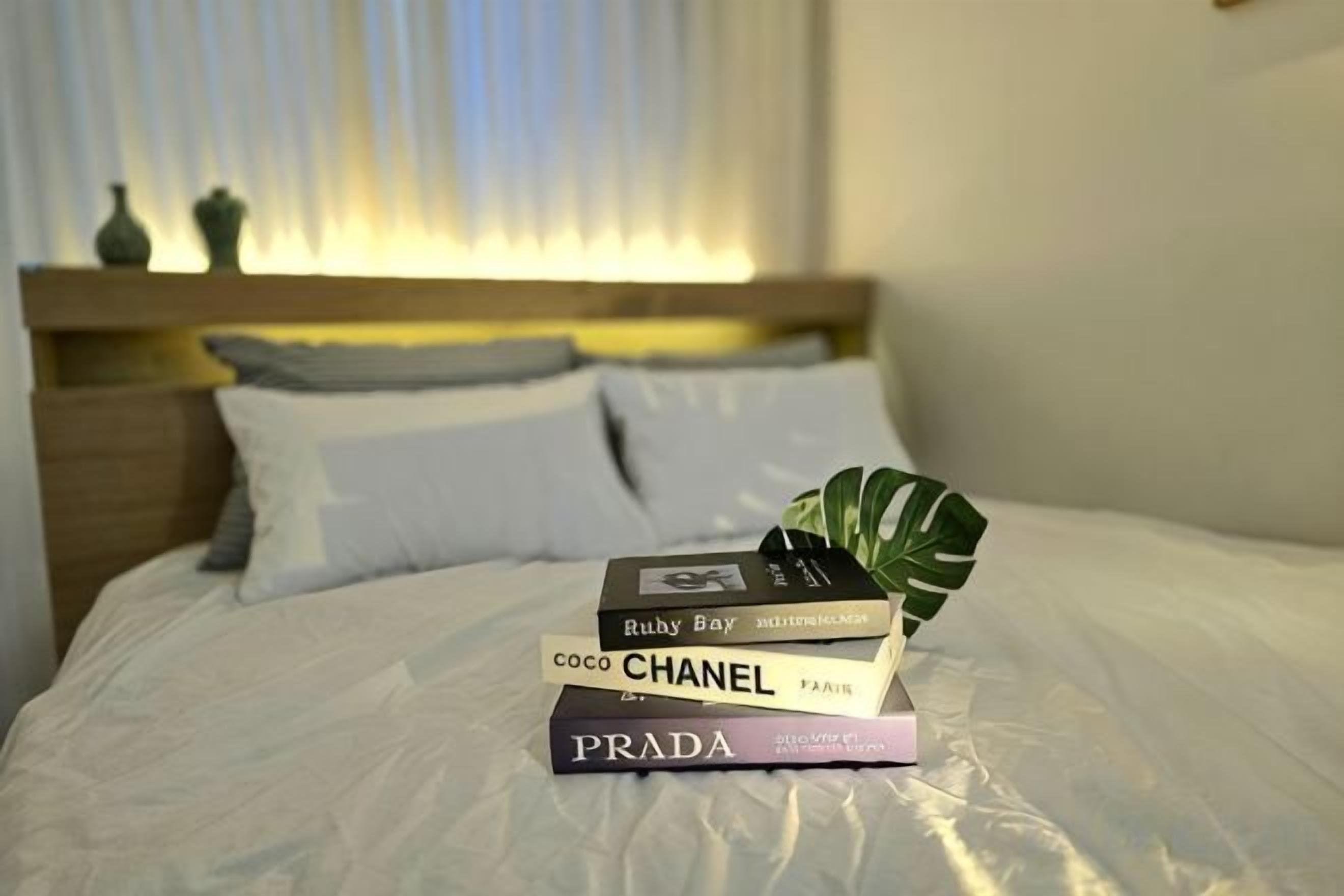 A neatly made bed with a stack of books on it, a potted plant, and a window with blinds in the background.