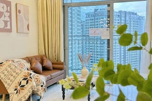 A living room with a brown sofa, a patterned throw, a small table with a vase, and a view of high-rise buildings through a glass door. - The Peninsula Apartment Landmark (Ho Chi Minh City)