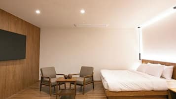 Un dormitorio moderno con suelo de madera, una cama grande con ropa blanca, dos sillones, una mesita y un televisor de pared.