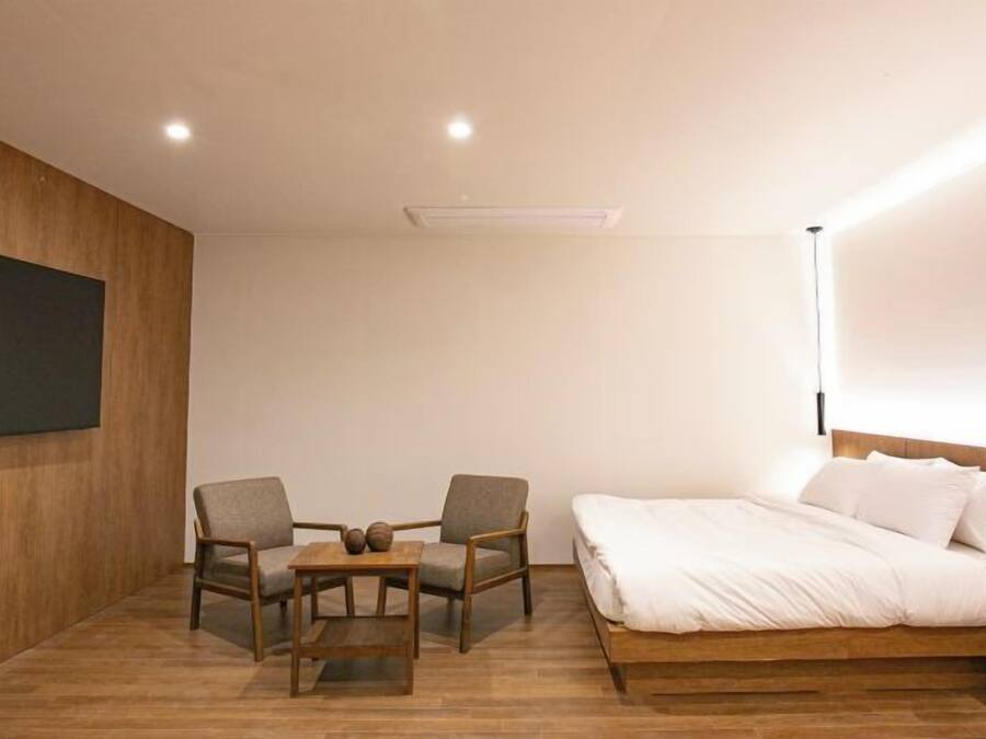A modern bedroom with a wooden floor, a large bed with white bedding, two armchairs, a small table, and a wall-mounted TV.