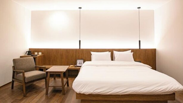 A hotel room with a wooden bed, a small table, a chair, and a nightstand with a lamp. - Hotel Emok (Busan)