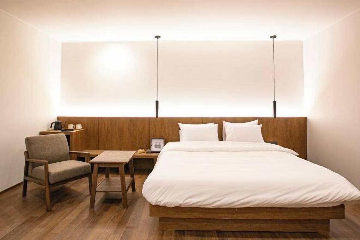 A hotel room with a wooden bed, a small table, a chair, and a nightstand with a lamp.