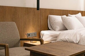 A hotel room with a wooden headboard, a bed with white linens, a grey armchair, and a wooden nightstand with a lamp and a small decorative item. - Hotel Emok (Busan)