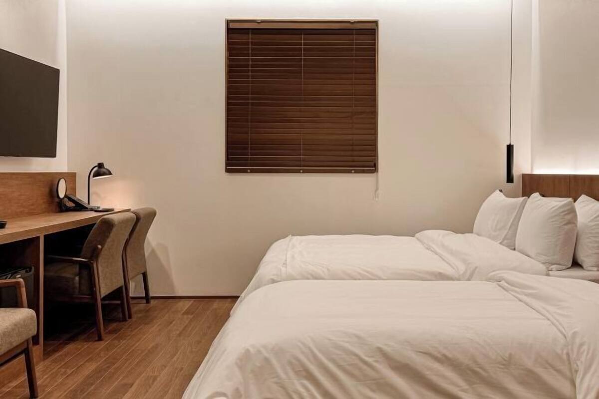 A hotel room with a bed, a desk with a chair, a lamp, and a window with blinds.