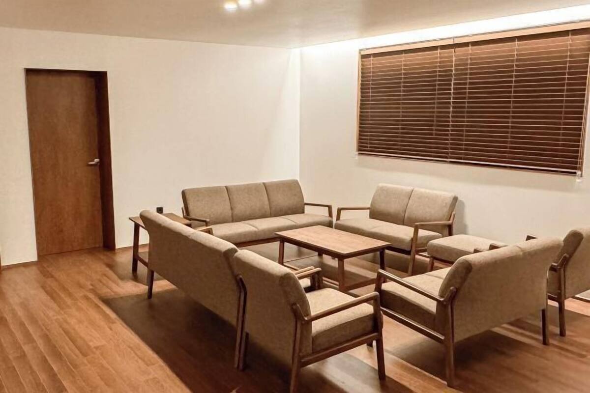A room with a sectional sofa, two armchairs, a small table, wooden flooring, a door, and a window with blinds.