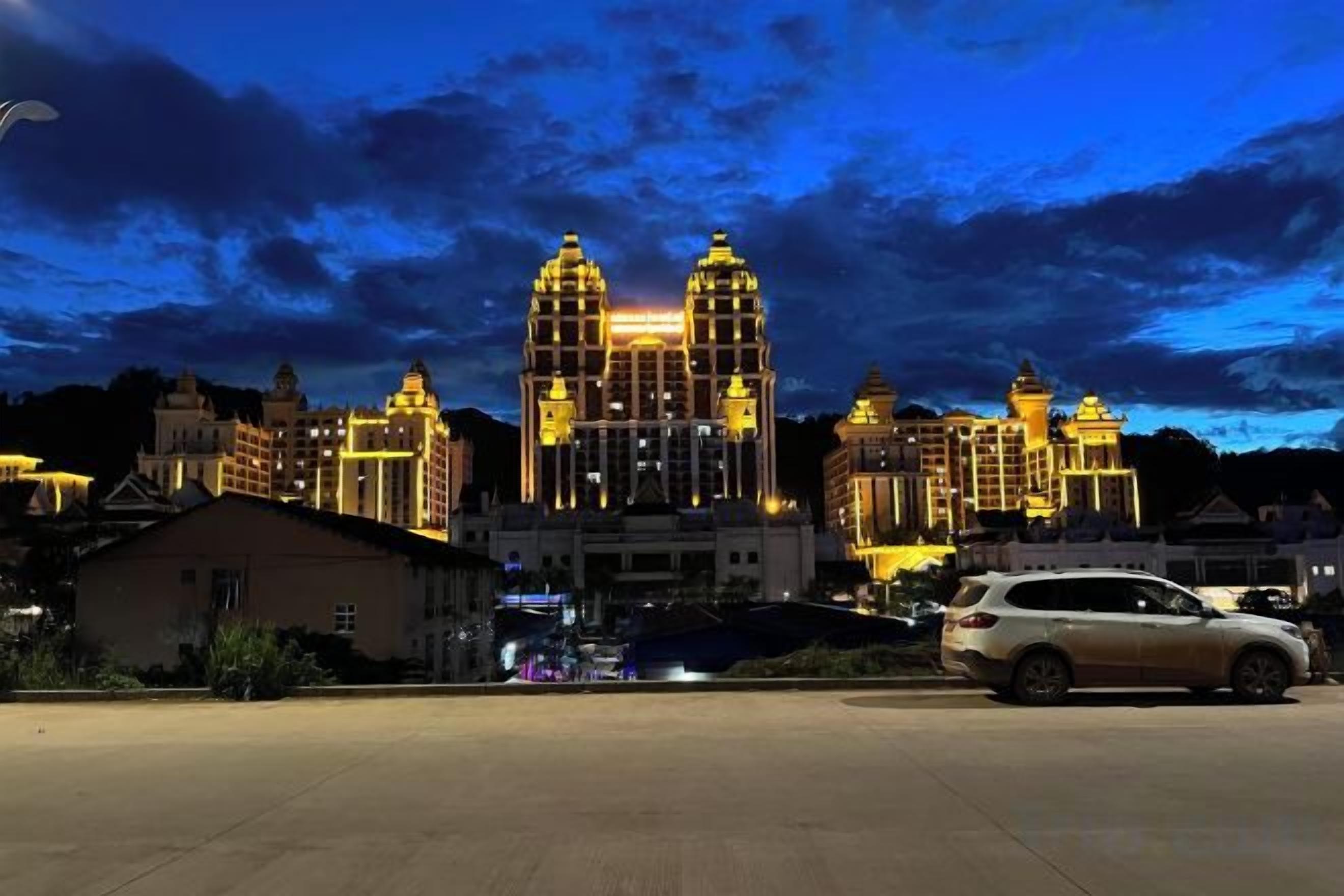 A large, illuminated building with multiple towers and a spacious parking area in front.