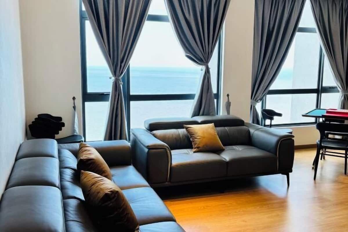 A modern living room with a sofa and armchair, large windows with curtains, and a view of the sea.