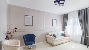 Un salon moderne avec un canapé beige, un fauteuil bleu et un fauteuil blanc. Deux œuvres d’art encadrées ornent le mur, ainsi qu’un lustre et une fenêtre avec des rideaux gris.