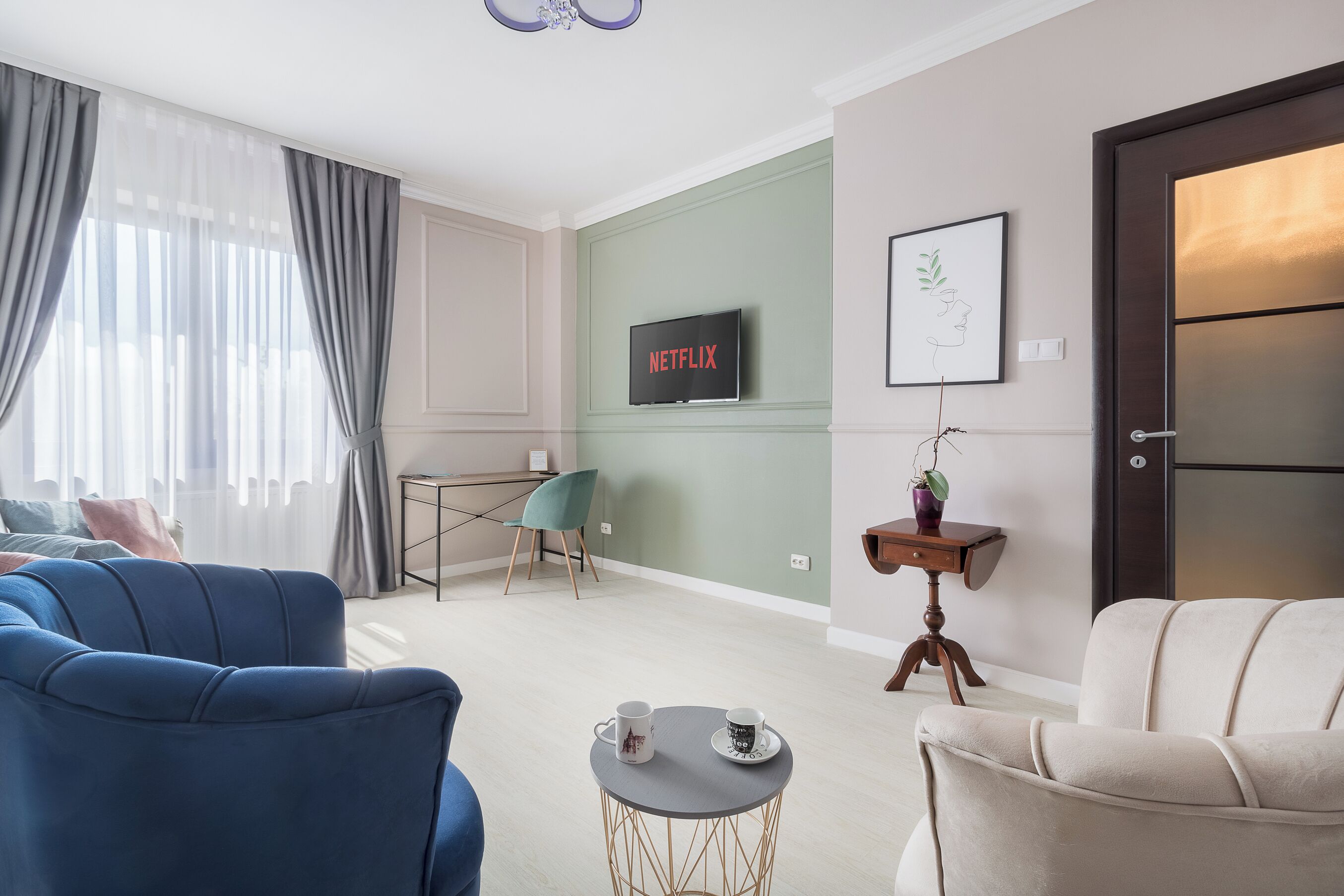 A modern living room with a sofa, armchair, and a small coffee table. A television is mounted on the wall, and there is a door with a frosted glass panel.
