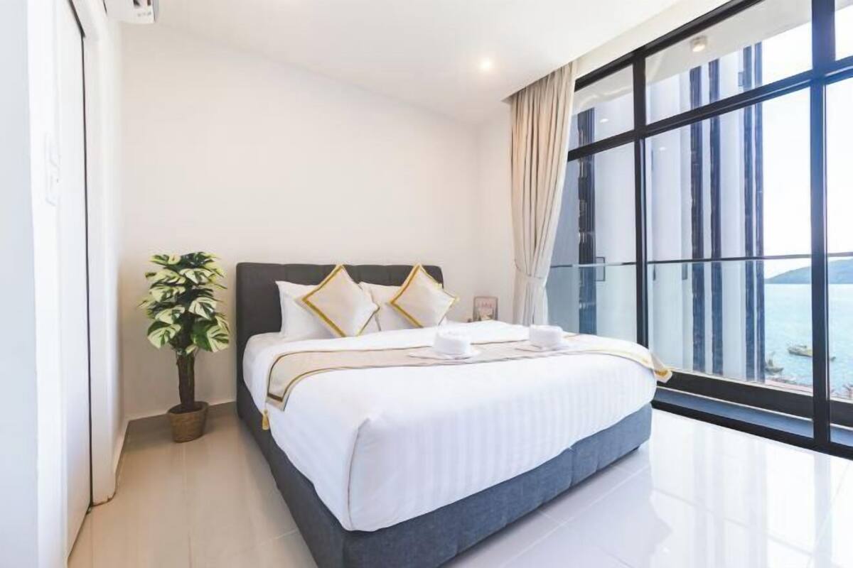 Une chambre moderne avec un grand lit, une plante en pot et une vue sur la mer grâce à de grandes fenêtres.