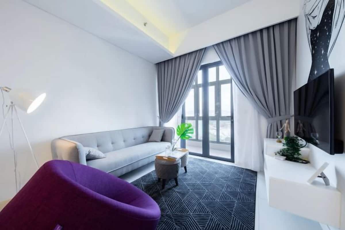Een moderne woonkamer met een grijze bank, een paarse fauteuil, een klein bijzettafeltje, een groot raam met gordijnen en een flatscreen televisie aan de muur.