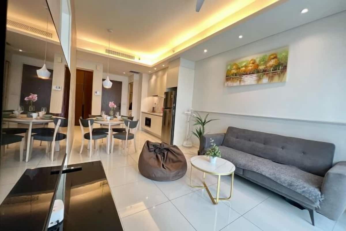 Een moderne woonkamer met een grijze bank, een eethoek met een tafel en stoelen, en een keuken op de achtergrond.