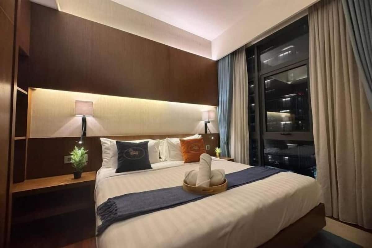 Hotelkamer met een groot bed, twee nachtkastjes met lampen, een bijzettafeltje, een potplant en uitzicht op de stad door een groot raam.