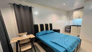 A compact room with a bed, a small desk, a chair, a nightstand, and a kitchenette area. - Cozy Studio Type unit w/ TV, Wifi, hotshower & ref (Cotabato)