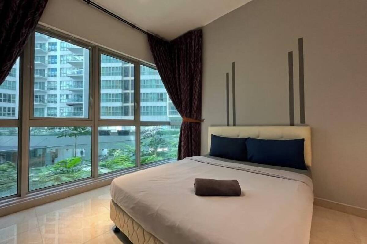 A hotel room with a large window, a bed with a white headboard, and a view of a building and greenery outside.