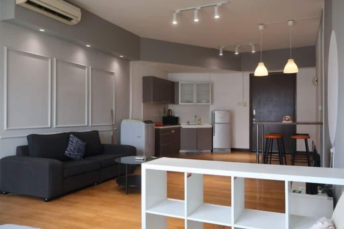 A modern living area with a sofa, a small table, a white shelving unit, a kitchen with a refrigerator, and a bar area with stools.
