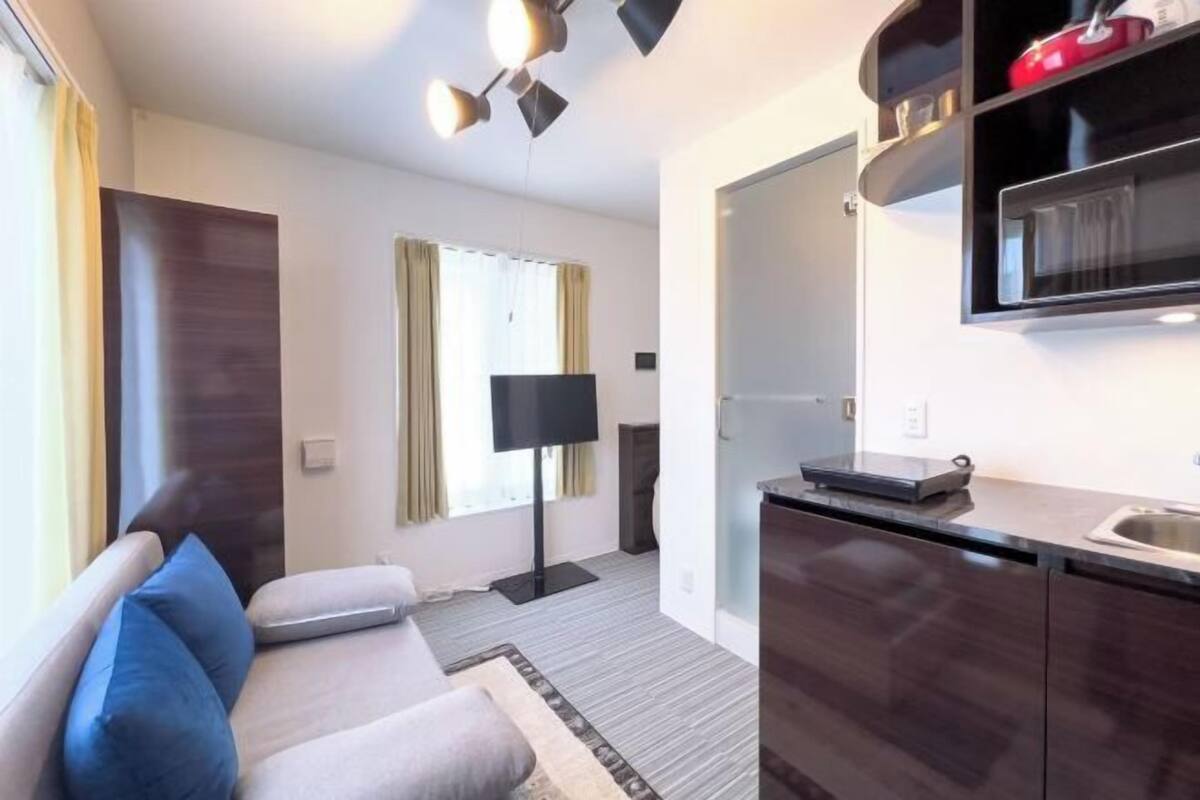 Un espacio habitable compacto con sofá, lámpara de pie, zona de cocina y puerta que da a otra habitación.