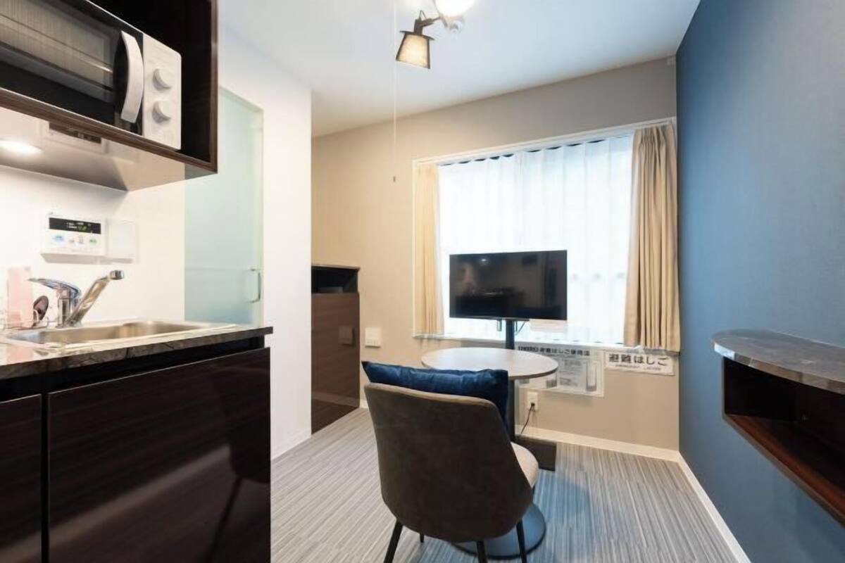 Habitación de hotel compacta con kitchenette, zona de comedor con mesa y silla, televisor de pantalla plana y ventana con cortinas.