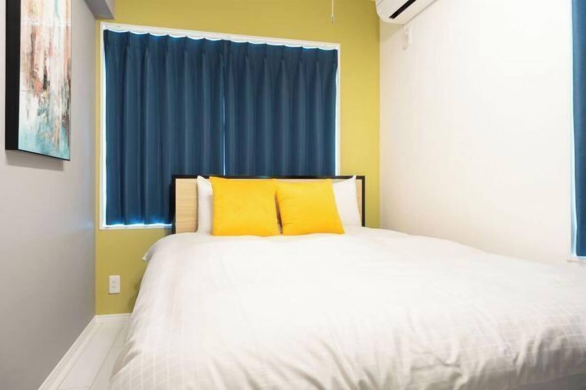 Una cama con ropa blanca y dos almohadas amarillas, una ventana con cortinas azules y un aire acondicionado fijado a la pared.