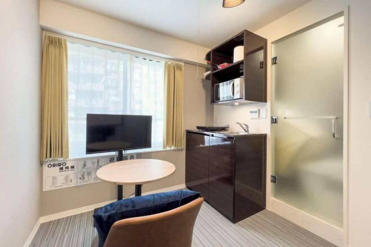 Habitación de hotel compacta con televisor, una pequeña mesa redonda, un sofá, kitchenette y una puerta de vidrio esmerilado.