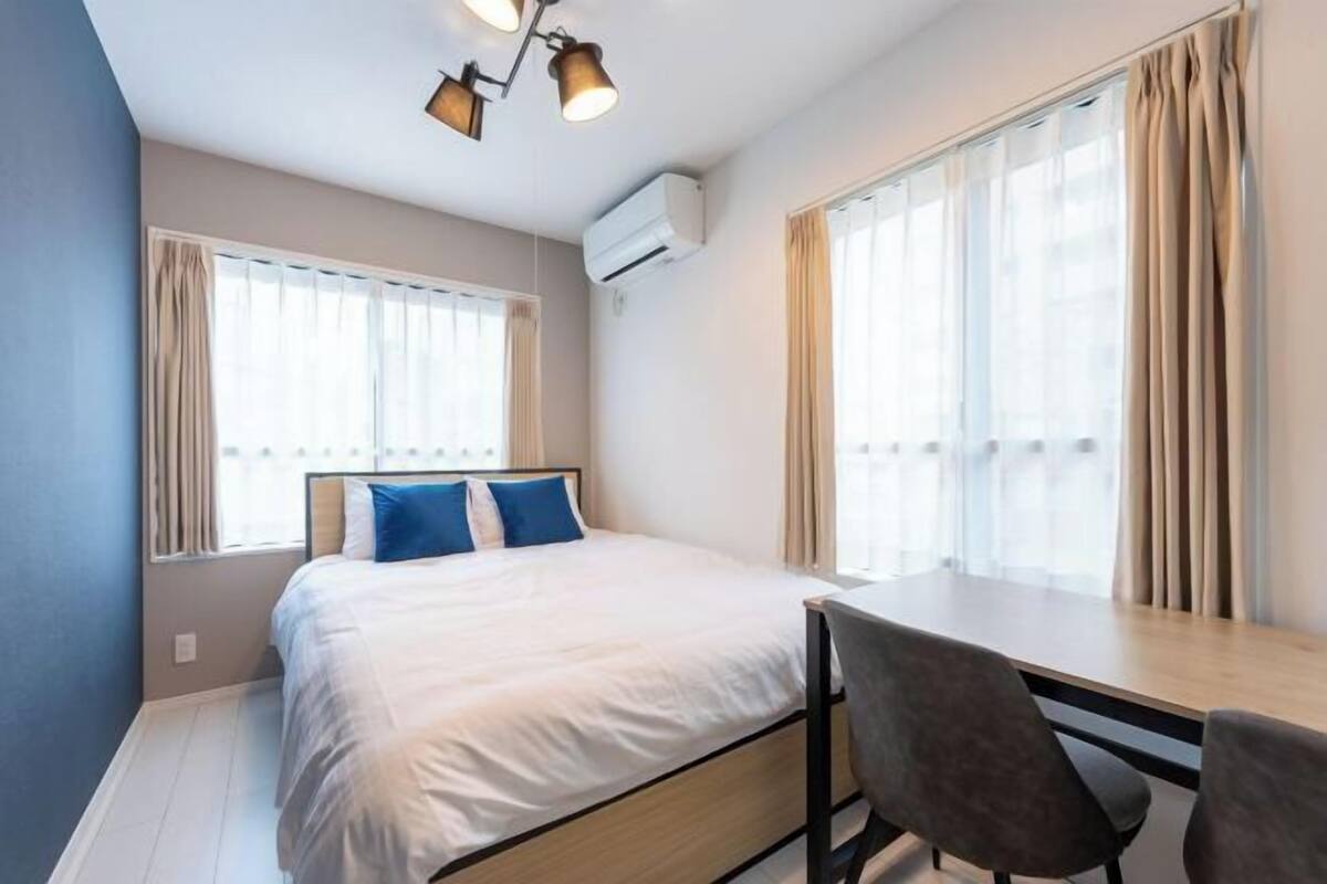 Un dormitorio con una cama, un escritorio y dos sillas. Hay ventanas con cortinas, un equipo de aire acondicionado y un aplique en el techo.