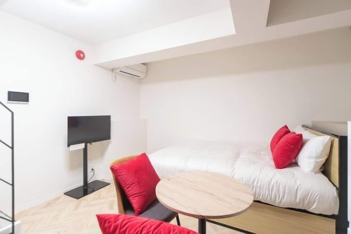 Una habitación pequeña con cama, una mesita redonda, un televisor y un cojín rojo.