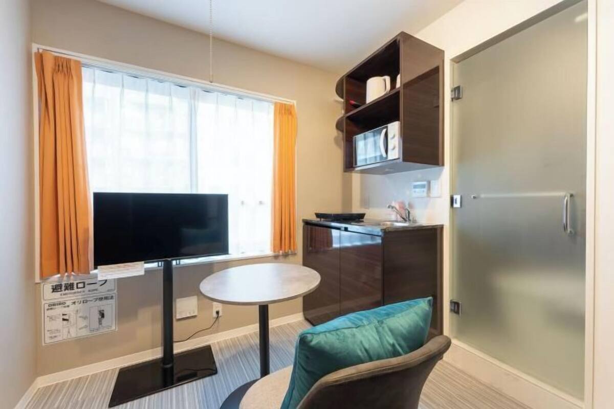 Una habitación de hotel compacta con una televisión de pantalla plana, una pequeña mesa redonda, una silla color aguamarina, una kitchenette y un armario con puerta corredera.