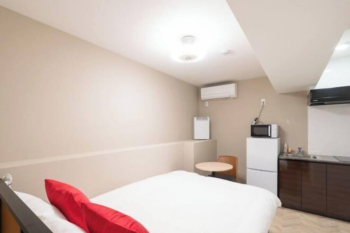 Una habitación de hotel pequeña con cama, una mesita redonda, kitchenette y una lámpara en el techo.