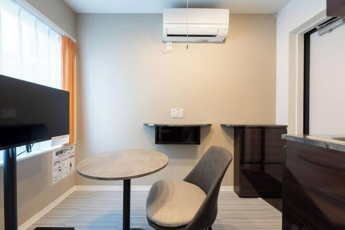 Una habitación compacta con televisión de pantalla plana, una mesa redonda, una silla acolchada, una pequeña zona de cocina con microondas y aire acondicionado integrado.