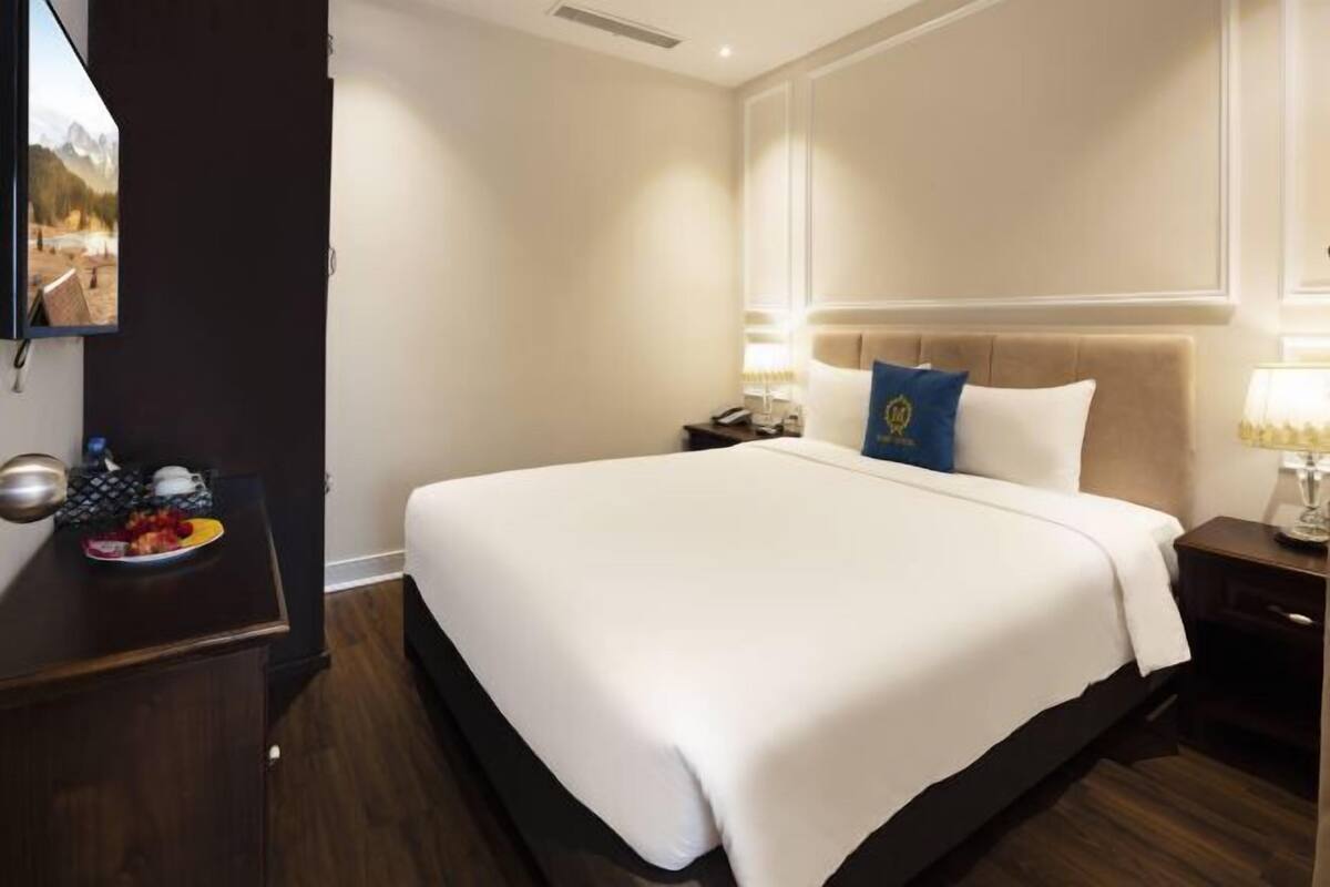 A hotel room with a large bed, a nightstand with a lamp, a small table with a plate of fruit, and a wall-mounted TV.